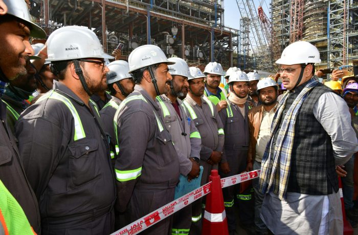 Construction workers in hard hats gathered on an industrial site for a briefing.